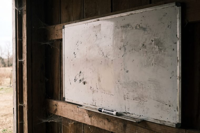 A small whiteboard screwed to a barn wall, wiped down to ghosted smears of old marker